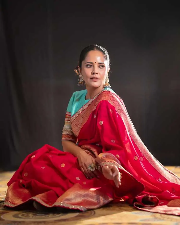 Anasuya Bharadwaj in a contemplative pose, seated on the floor, captivating in her traditional red silk saree.