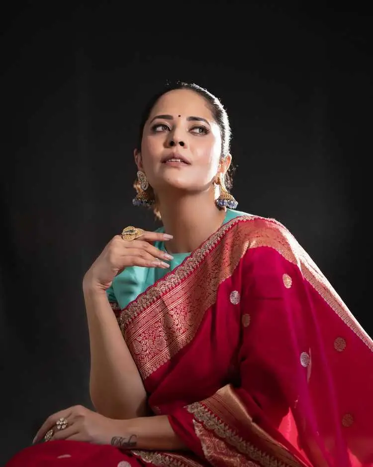 Anasuya Bharadwaj looking elegantly away, draped in a beautiful red and gold silk saree, with traditional jhumkas.