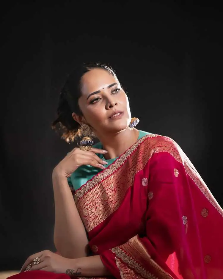 Anasuya Bharadwaj posing gracefully, showcasing her stunning red silk saree and intricate jewelry against a dark backdrop.